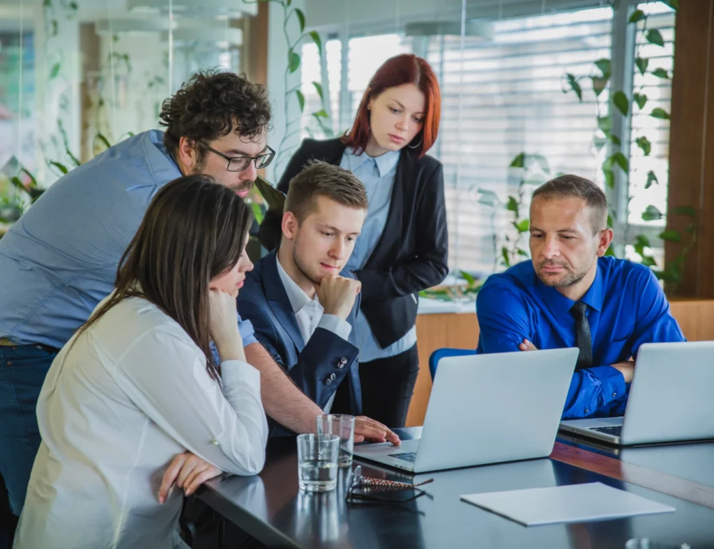 A group of people sitting around a table in an office building, looking at a laptop. They are smiling and engaged in discussion, with a window visible in the background.