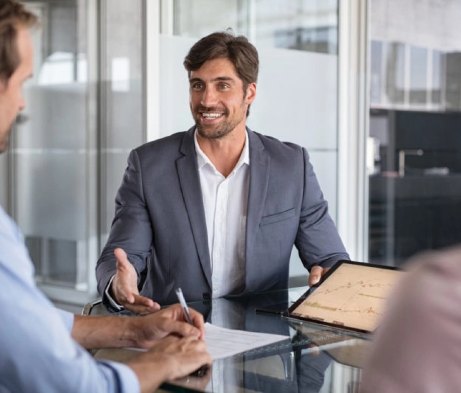 Man in suit sitting at a glass table showing a graph on a tablet to two people while explaining something in a modern office setting.